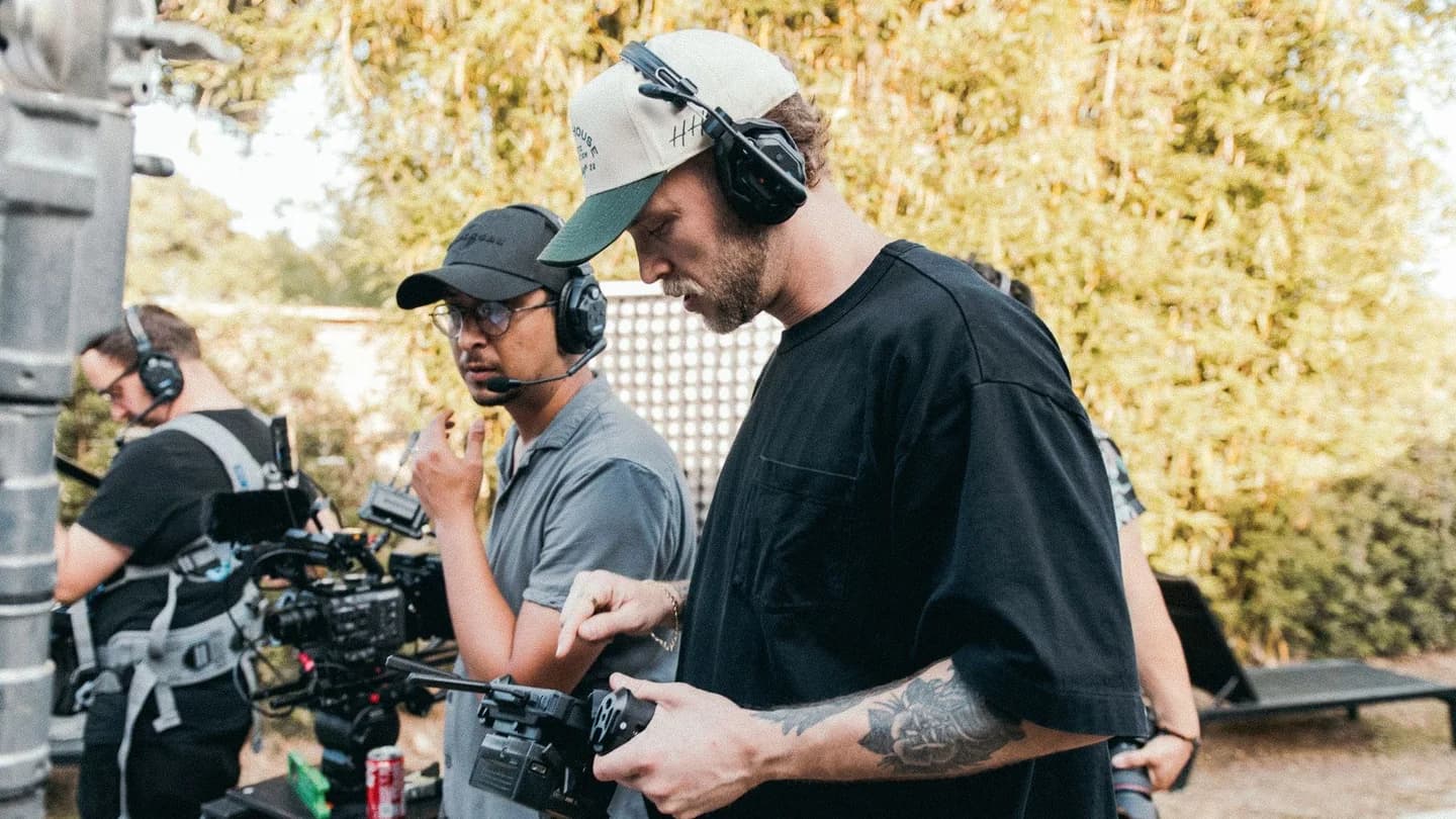 An outdoor daylight film set: two crew members in caps and headsets stand at a camera rig, one working the lens controls, a third crew member operating a camera behind them, foliage in the background.